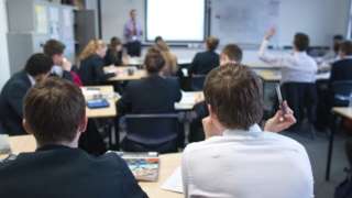Stock image inside a classroom. Students can be seen sitting at desks whilst a teacher stands at the front by a whiteboard.