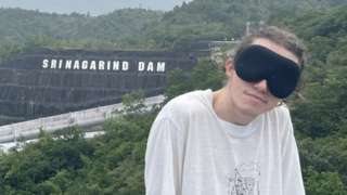 Ed Smith, blindfolded, standing beside view of dam in Thailand
