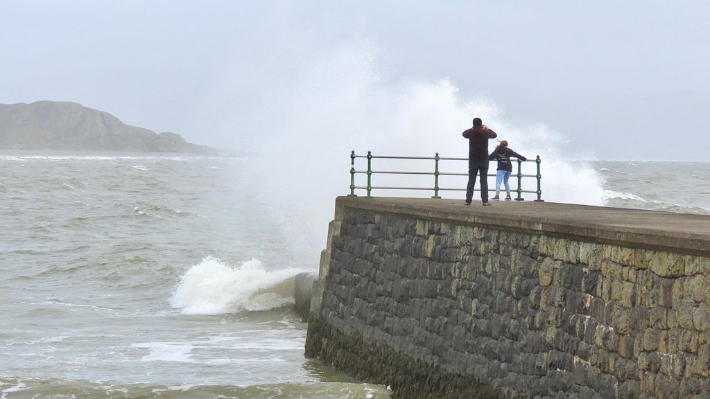 Two people watch the stormy seas at the end of a pier in Criccieth, Wales