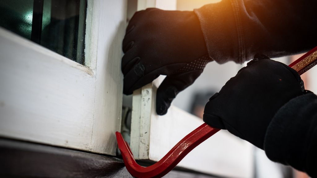 A stock image showing a person in gloves using a crowbar to open a window to gain access to a home
