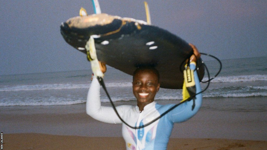 A smiling Vanessa Turkson walks out of the surf carrying a board on her head