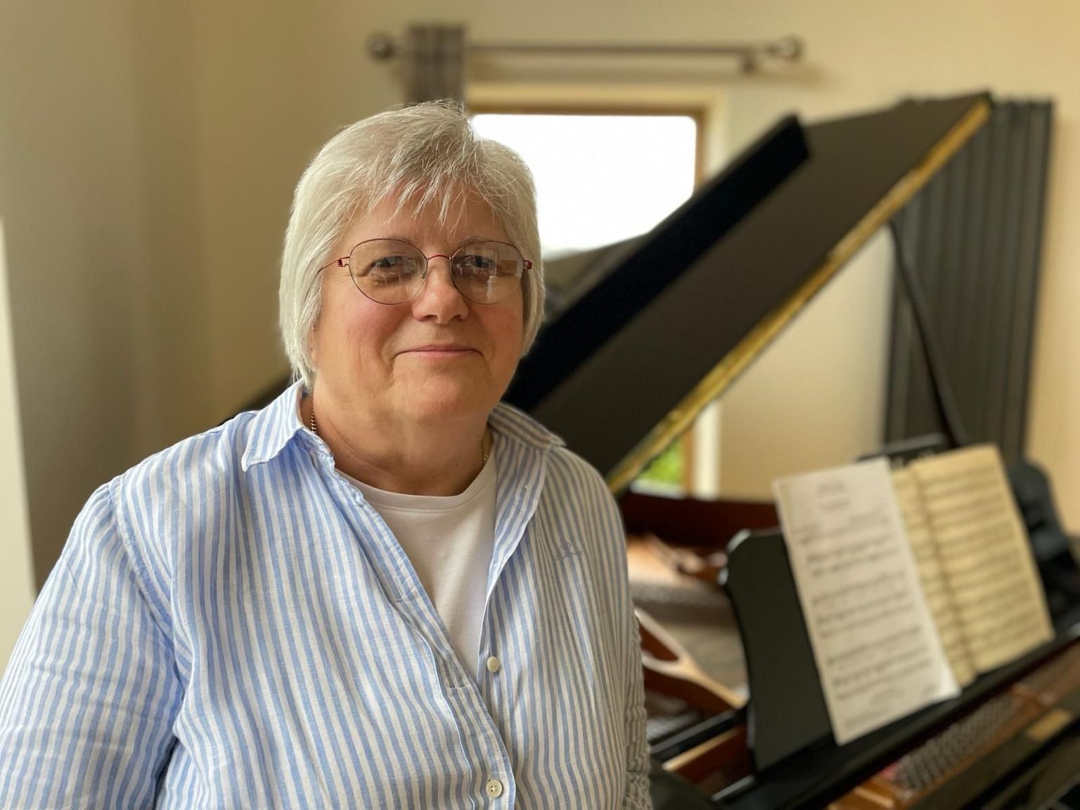 Ann is a white woman with short, grey hair and she is wearing glasses. She is stood next to her late husband's grand piano.
