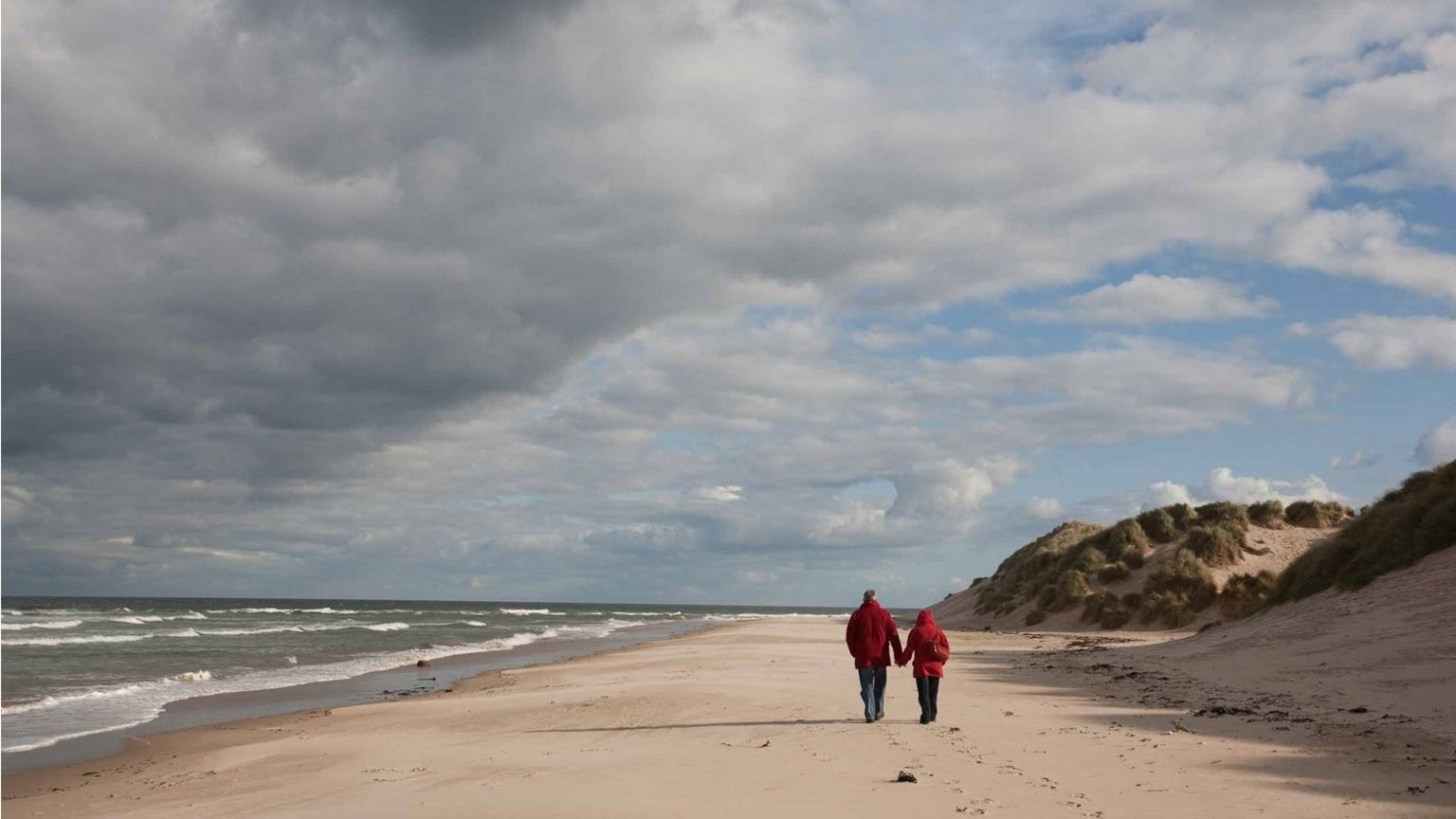 Two people walk hand in hand on a soft white, wild and natural, beach with dunes