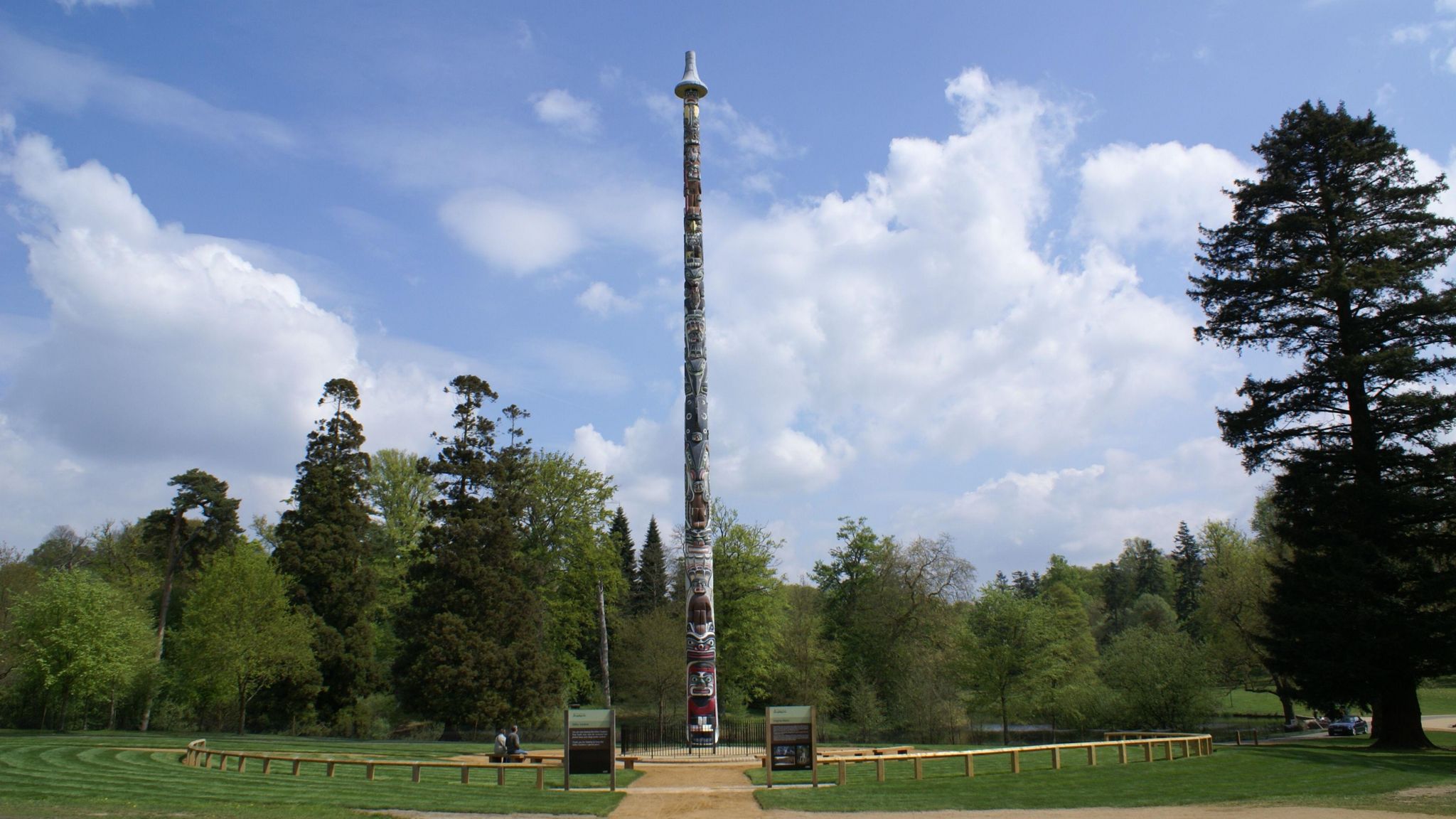 A totem pole stands in the middle of the image. There are trees around it and a low fence