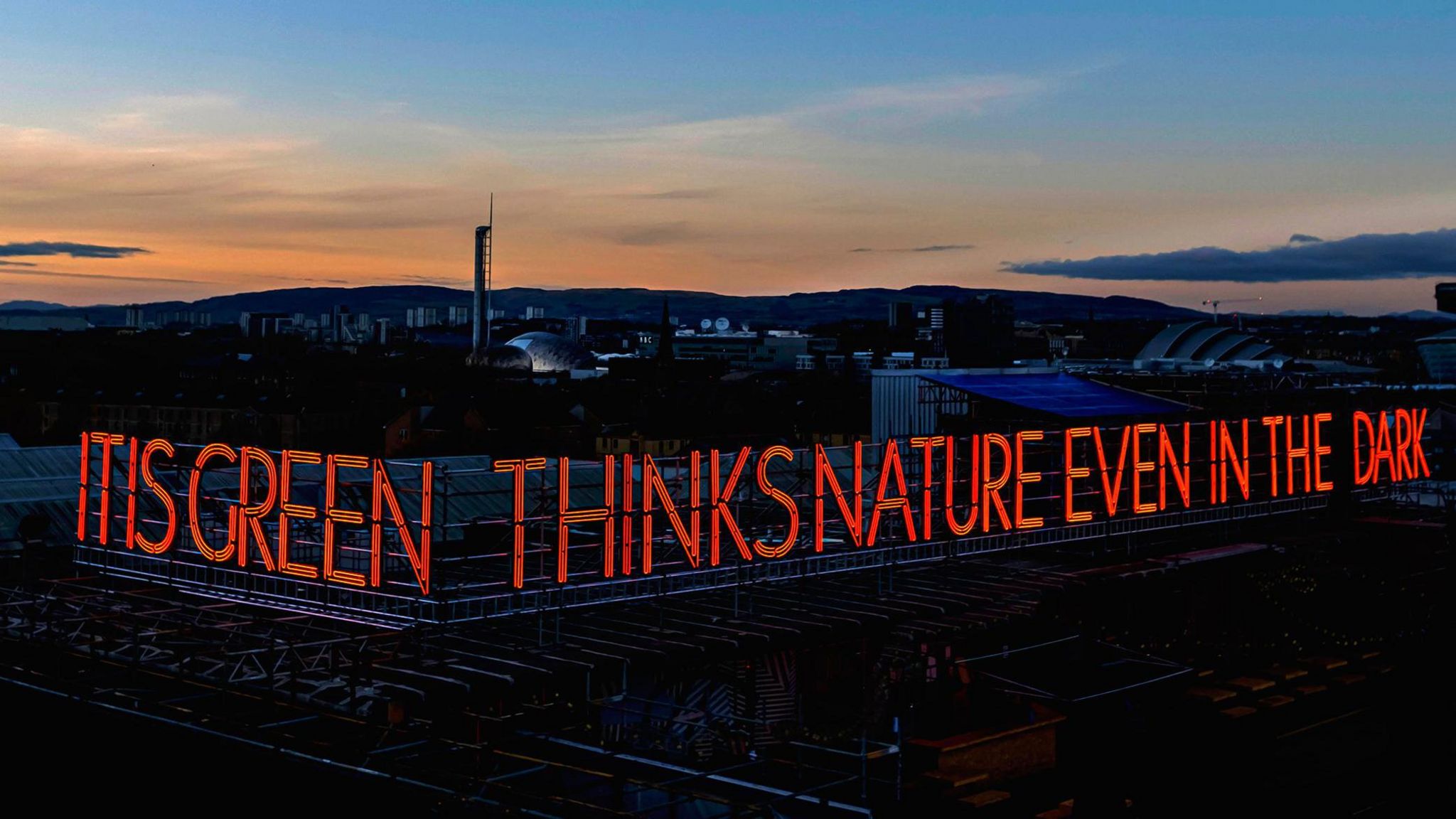 A photo of the neon sign reading "IT IS GREEN THINKS NATURE EVEN IN THE DARK" with a sunset in the background