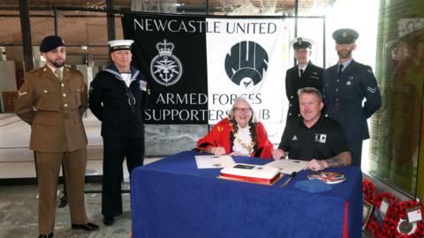 Forces veterans stood in front of the clubs banner, with the Lord Mayor smiling at a table with a blue tablecloth.