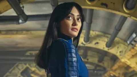 A young woman with long black hair stands in an artificially lit, cylindrical corridor with various pipes supported by bright yellow struts along its length She's looking back over her shoulder with an impassive expression. She wears a close-fitting, dark blue leather jumpsuit with panelling down the arms.