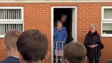 A woman standing in the doorway of her house as people sing carols outside