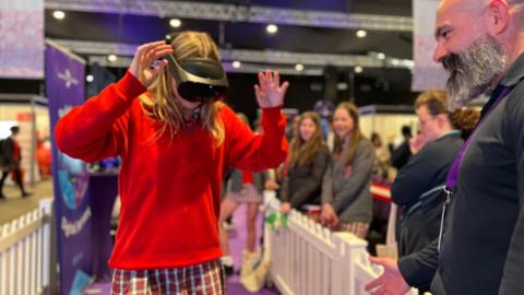 A female student in a red jumper wears a virtual reality headest  with her hands in the air as a man from Digital Jersey gives her advice