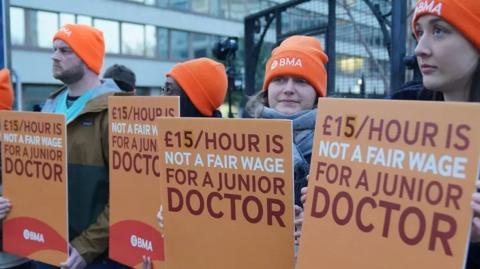 Doctors manning a picket line carrying signs saying '£15 an hour is not a fair wage for a junior doctor'
