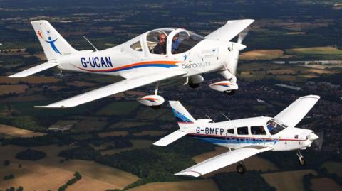 Two small white aircraft with Aerobility written along the side fly side-by-side above patchwork fields