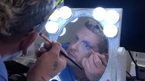GP Duncan applying eye shadow in the mirror, surrounded by lights