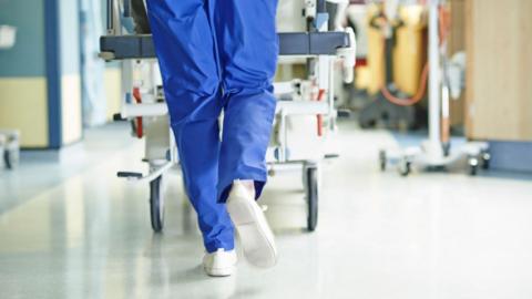 A medical staff member in blue scrubs, we see only the back of their legs as they push a trolley