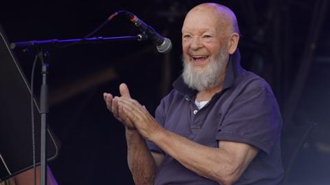 Image of Michael Eavis. He is pictured behind a microphone, smiling and clapping his hands.