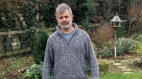 Kevin Lister, with grey hair and beard, stands in a garden wearing a grey knitted jumper