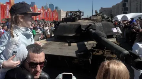 A little girl on a man's shoulders next to a tank