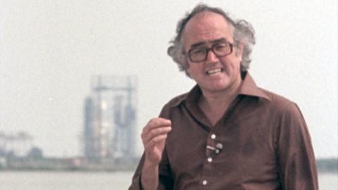 James Burke giving an explanation to camera. In the background is a rocket about to take off. 