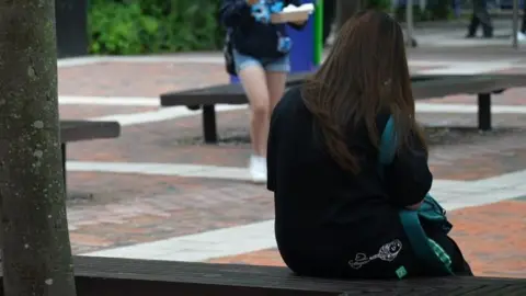 Image of an anonymous student sitting on a bench in a square on a university campus.