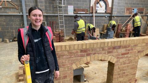 Woman wearing black jacket and pink high viz jacket stands in front on an arch brick wall she built