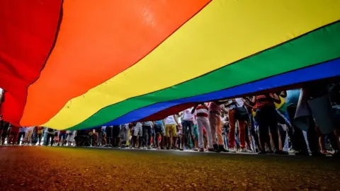 A camera shot taken from beneath a large LGBT rainbow flag shows people's legs standing to the right of the flag