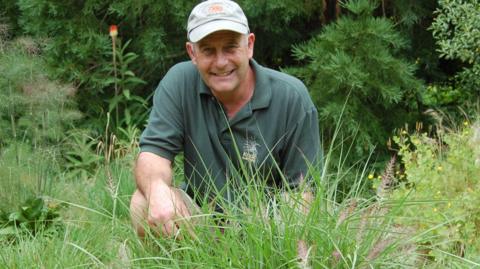Neil Lucas wearing white cap and crouching among the ornamental grasses