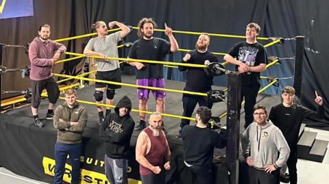 Men in casual sports clothes post next to a wrestling ring.