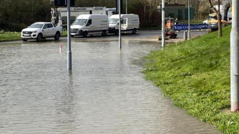 A flooded road with work vans at the edge and lampposts and a cone coming through the water