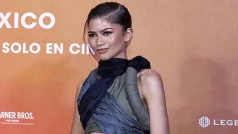 Zendaya wearing one of Torishéju Dumi's designs at a Dune premiere in Mexico City in February. Zendaya is a 27-year-old actress with brown eyes and dark hair which is slicked back with a severe side parting. The dress is high necked and made up of three different materials - blue, silver and green - draped in bands across her chest.