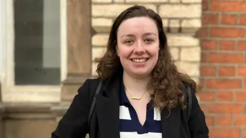 Nichole Brown. Nichole is a 25-year-old white woman with long curly brown hair. She has blue eyes and smiles at the camera. She wears a black coat over a blue and white striped top with a gold necklace and a black back pack on her shoulders. She's pictured outside in Stoke-on-Trent in front of a brick wall