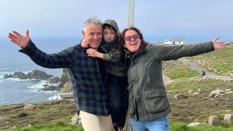 Caroline with her husband Carlos and their son, standing with their arms outstretched at the Land's End sign in Cornwall