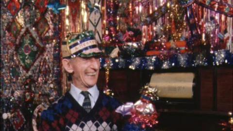 'Tinsel Man' Fred English smiling in his tinsel-covered living roon, wearing a hat with tinsel on it.