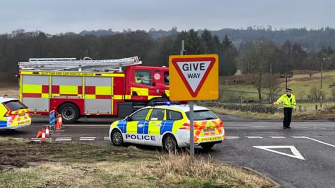Two police cars and a fire engine with a police officer standing on the road - a t-junction among fields at a give way sign