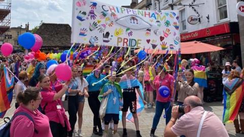 Chippenham Pride 2023 with people surrounded by multi-coloured balloons and flags with a group in the centre of the picture carrying a pride banner