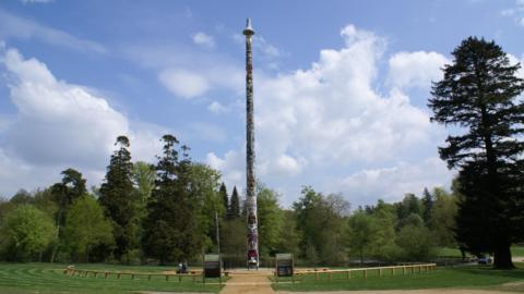 A totem pole stands in the middle of the image. There are trees around it and a low fence