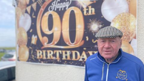 Man in a flat cap stood in front of sign that reads 'Happy 90th birthday'