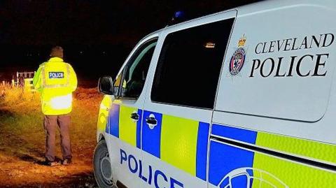 A police officer in a rural location at night, standing next to a police vehicle