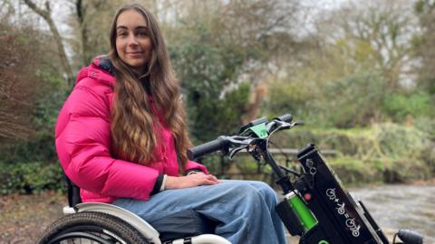 Bethany Handley sitting in her wheelchair with a power attachment looks at the camera while outdoors