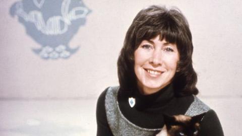 Valerie Singleton sitting in the Blue Peter studio. She's holding Jason the cat, but only the top of his head is visible.