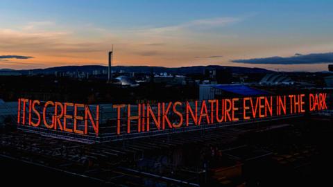 A photo of the neon sign reading "IT IS GREEN THINKS NATURE EVEN IN THE DARK" with a sunset in the background