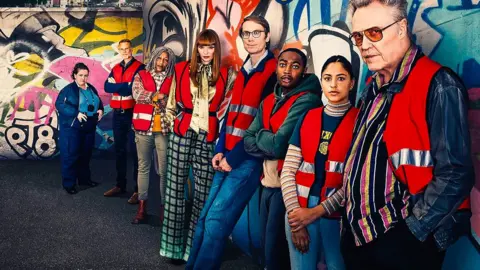 The cast of The Outlaws lined up against a graffitied wall, wearing red hi-vis vests