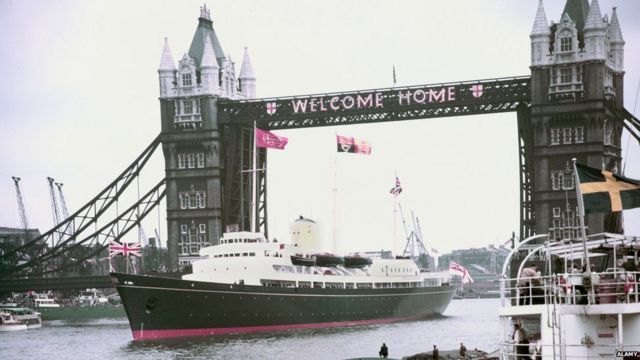 Britannia проходит под Тауэрским мостом, на котором вывешено приветствие: "Добро пожаловать домой"