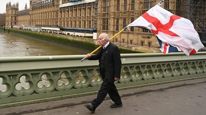 Мужчина с флагами Англии и Британии около здания британского парламента.