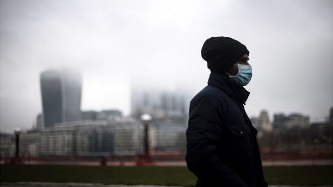 Man walks along south bank of Thames through city of London on a grey day, with a mask on