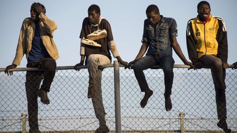 Four African migrant men sit on a mesh boarder fence separating Morocco from the Spanish enclave of Melilla