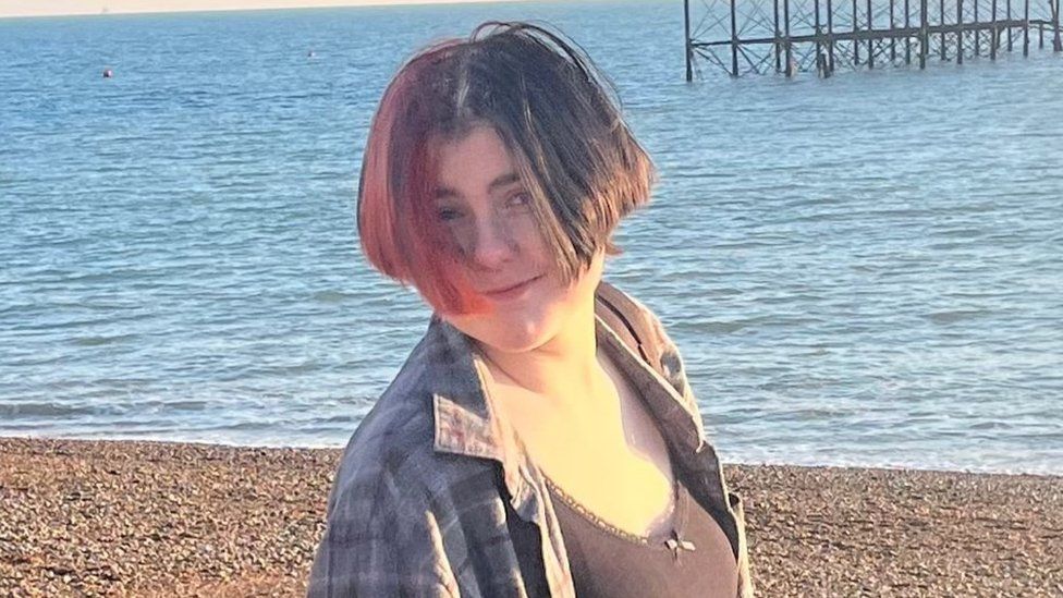 EJ stands on a pebble beach on a sunny day with the shoreline and sea visible in the background. Part of the metal frame of an old pier can also be seen. EJ's hair - which is half-brunette, half-red, partly covers EJ's smiling face. EJ wears a black vest top with an unbuttoned grey/black checked shirt over the top.