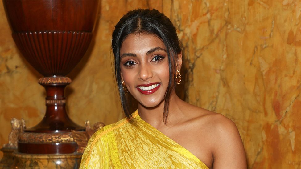 Charithra Chandran. Charithra is a 27-year-old British Asian woman and is pictured smiling at the camera. She has brown eyes and long dark hair which she wears tied back with a long fringe framing her face. She wears a one-shouldered yellow dress and gold hoop earrings. Her lips are painted red and she wears black eyeliner. Charithra is pictured inside against a gold marble wall with a brown ceramic vase.