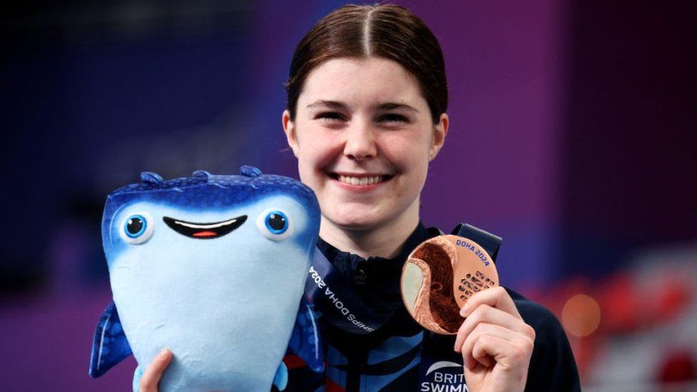 Bronze Medalist, Andrea Spendolini Sirieix of Team Great Britain pose with their medals during the Medal Ceremony after the Women's 10m Platform Final on day four of the Doha 2024 World Aquatics Championships. Andrea is a 19-year-old woman with dark brown hair tied back. She smiles at the camera as she holds up a bronze medal and a plush smiling stingray mascot. She wears a navy blue British Swimming tracksuit.
