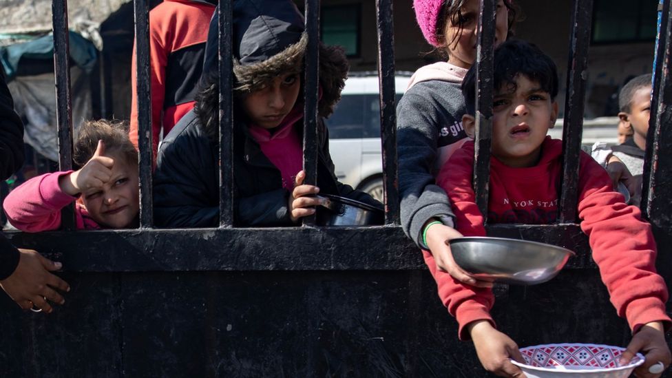 Displaced Palestinian children hold empty pots as they line up to receive food aid provided by a Palestinian youth group, at Rafah refugee camp