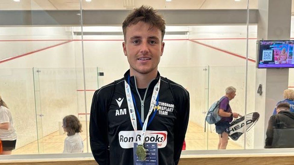 A young, white man wearing a black sports top with a gold medal stands in front of squash courts. He's got short, slightly dishevelled hair and is wearing a medal around his neck on a white lanyard. The squash courts are behind a glass screen - they're mostly a bright white colour, apart from the red lines denoting the area of play.