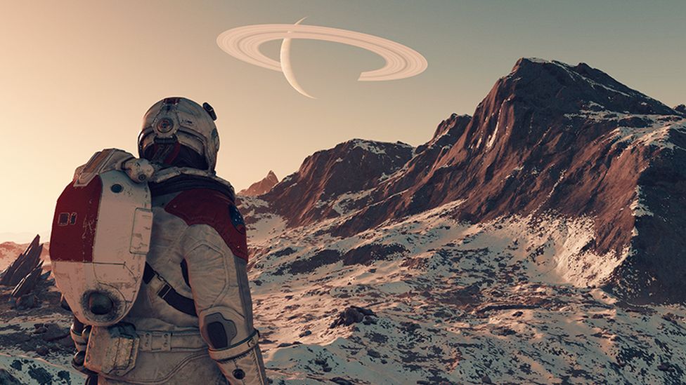 A screenshot from Starfield shows the back of an astronaut character in spacesuit and helmet surveying a barren planetscape. It is rocky, with cliffs jutting out of the ground. The rocks are covered with a white powder - either dust or snow. In the distance another planet, and its horizontal rings, can be see in the sky.